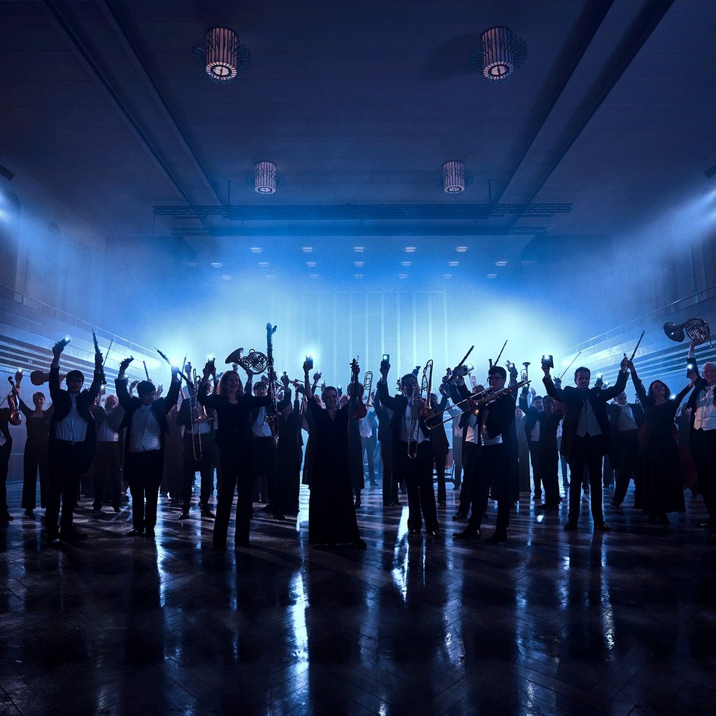 Orchester im dunklen Konzerthaus, nur Schatten der Personen sind sichtbar. Alle strecken die Hände, teilweise mit Instrumenten in der Hand, in die Luft. 