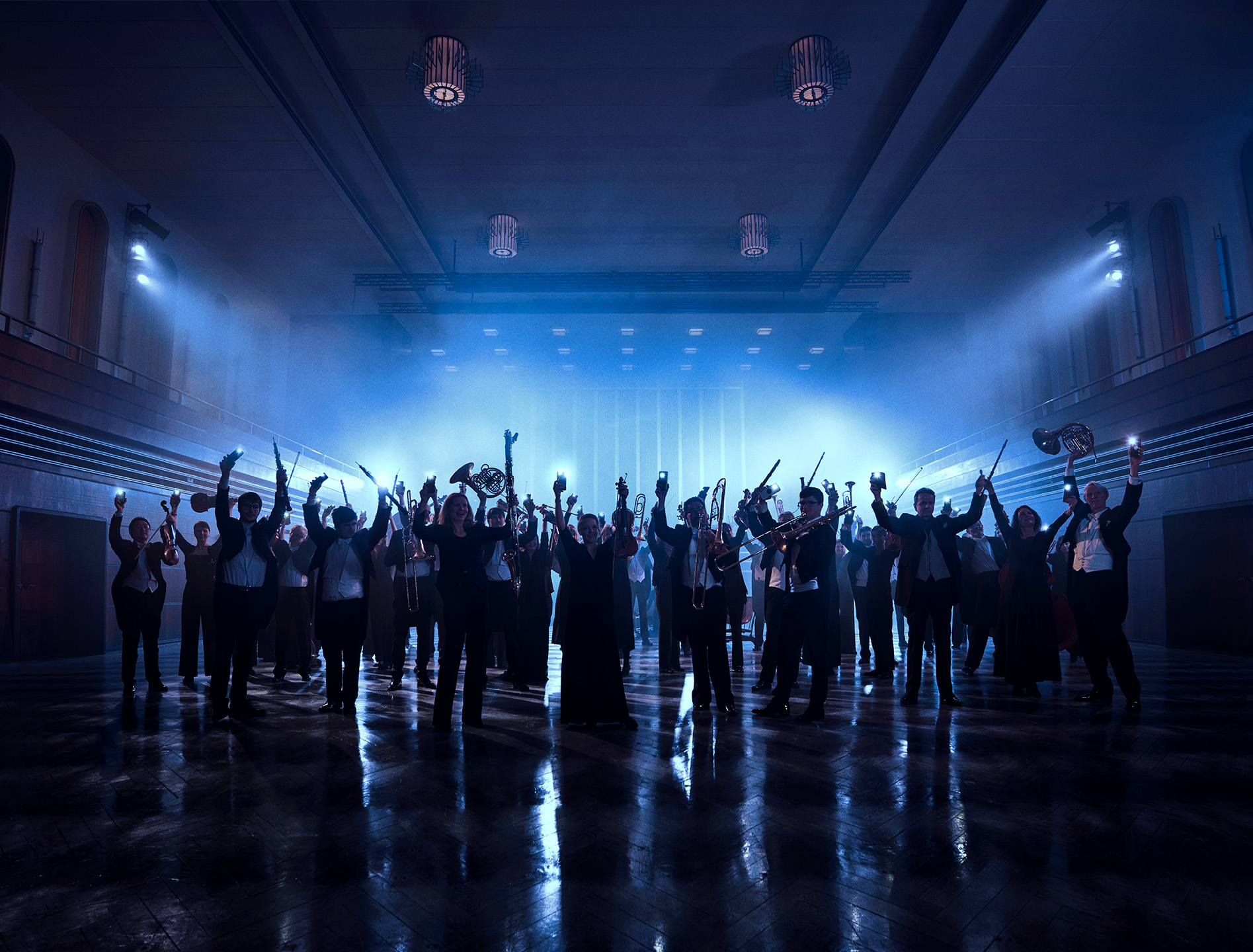 Orchester im dunklen Konzerthaus, nur Schatten der Personen sind sichtbar. Alle strecken die Hände, teilweise mit Instrumenten in der Hand, in die Luft. 